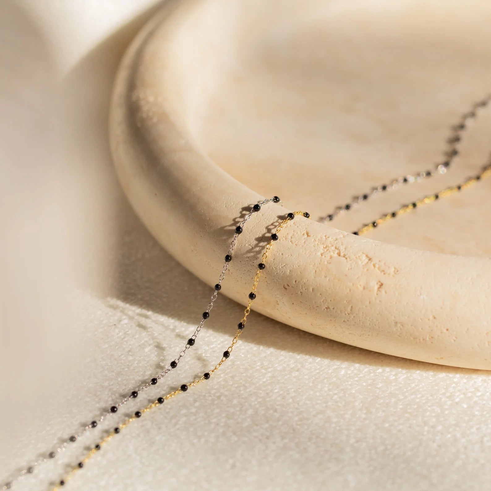 A close-up of the Onyx Necklace Set, a delicate gold and black chain perfect for layering, draped over a round cream-colored ceramic plate.