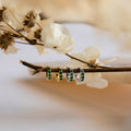 The Elise Gemstone Huggies Set, featuring four small artisan-made earrings with green and yellow stones, is displayed on a branch accented with dried white flowers.