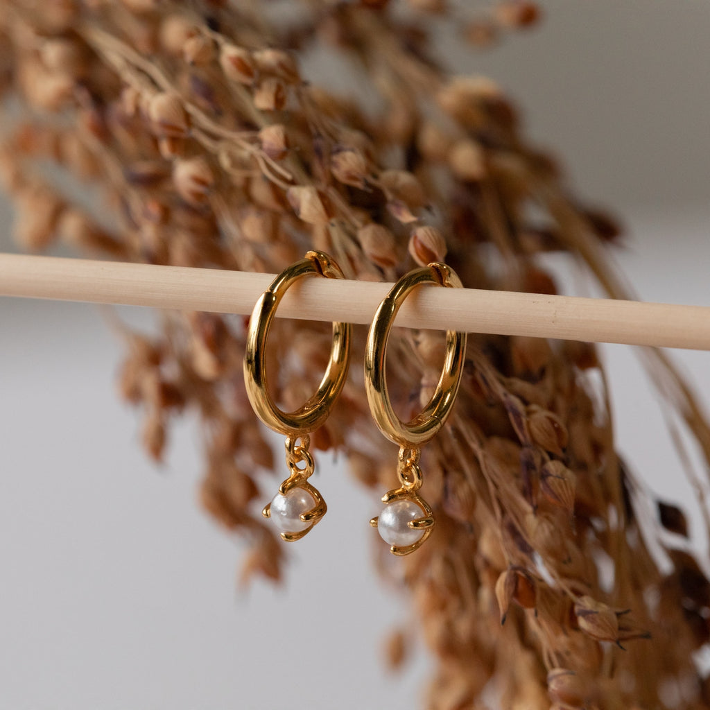 Molly Pearl Huggies in Sterling Silver feature small dangling pearls and elegant hoops, displayed on a wooden rod with softly blurred dried grass in the background—perfect for adding timeless charm to any look.