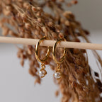 Molly Pearl Huggies in Sterling Silver feature small dangling pearls and elegant hoops, displayed on a wooden rod with softly blurred dried grass in the background—perfect for adding timeless charm to any look.