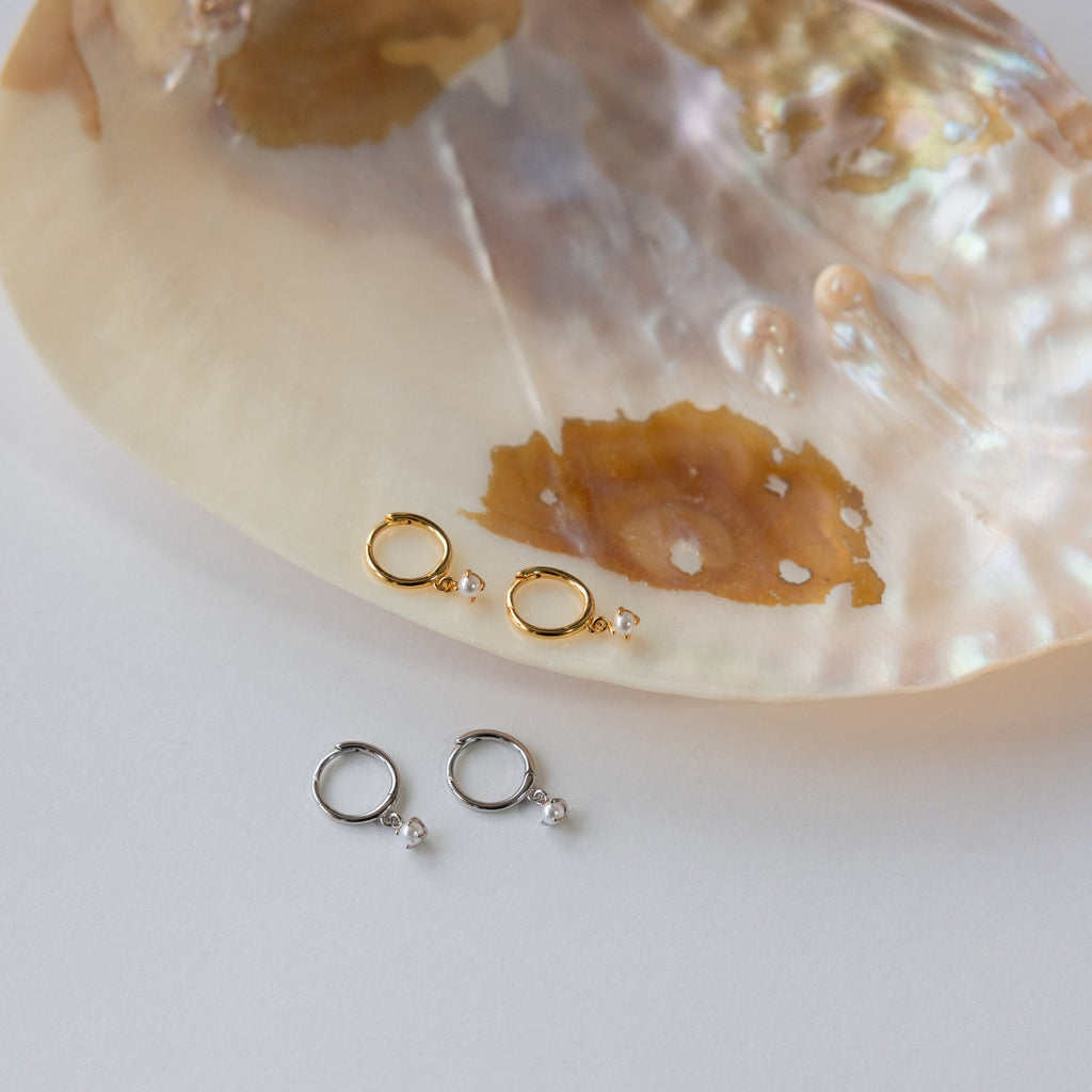 Two pairs of Molly Pearl Huggies, one gold and one silver, displayed on a large iridescent seashell.