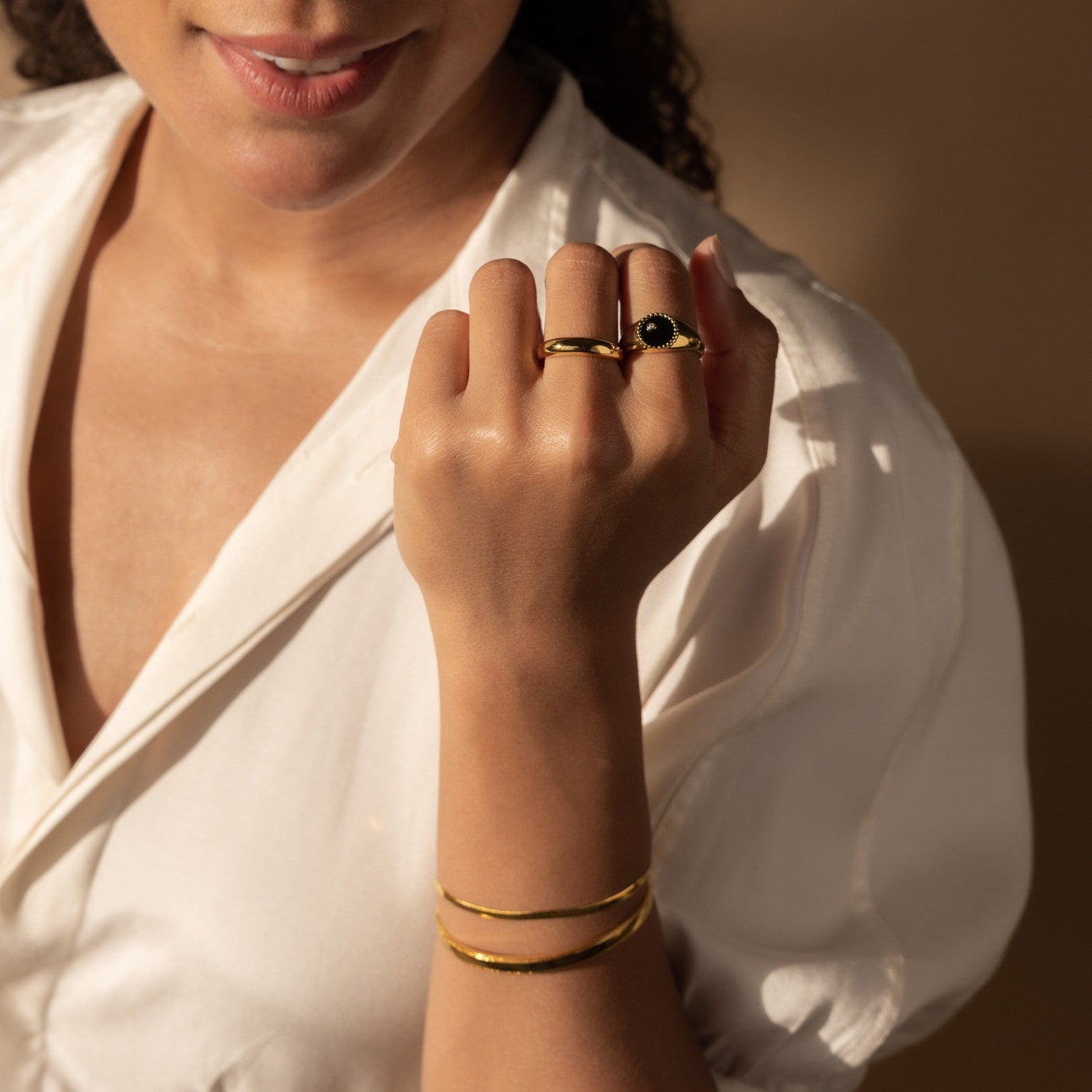 Woman in white blouse displays gold jewelry, including bracelets and the Natasha Black Signet Ring, with sunlight casting beautiful shadows.