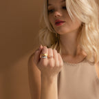 A blonde woman displays the Carrie Signet Ring alongside minimalist gold bands on her fingers, set against a beige background.
