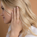 A woman with blonde hair touches her ear, wearing the Initial Morse Code Ring on her finger—a stunning personalized gold ring accented with sparkling stones.