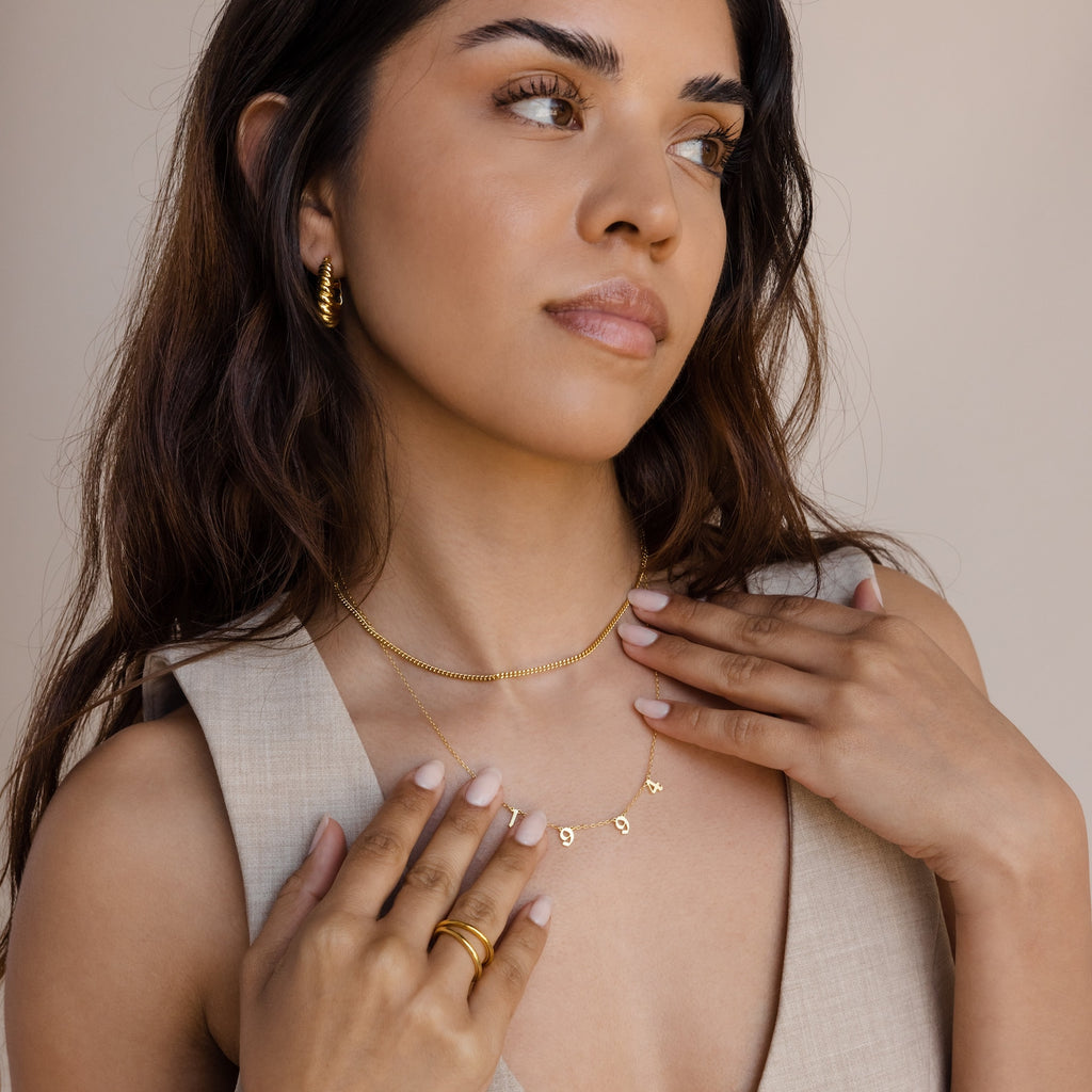 A woman wears gold jewelry—hoop earrings, rings, and a vintage charm necklace layered with the Old English Spaced Number Necklace—against a neutral background.