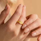 A hand with nude nails wears a minimalist gold Old English Signet Ring engraved with the numbers 444 on the index finger.