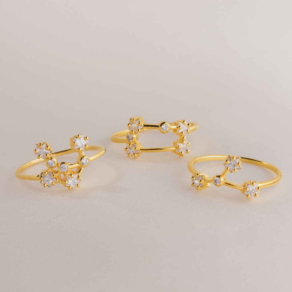 Three delicate gold Constellation Rings with small, sparkling flower-shaped designs evoke celestial jewelry, beautifully displayed on a light surface.