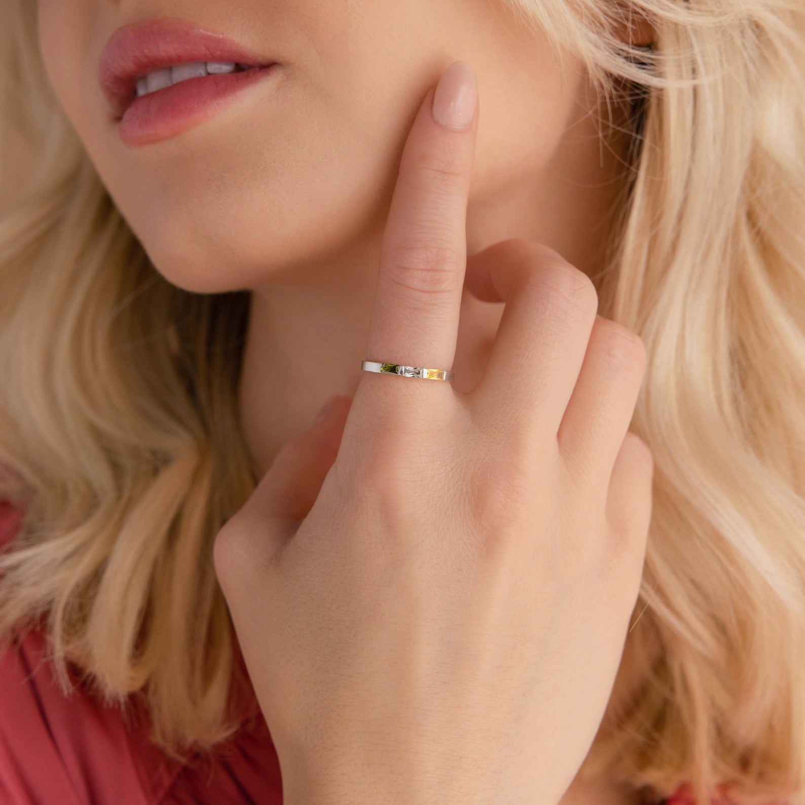 A woman with blonde hair touches her face, wearing the Mothers Baguette Birthstone Ring on her finger and a pink top.