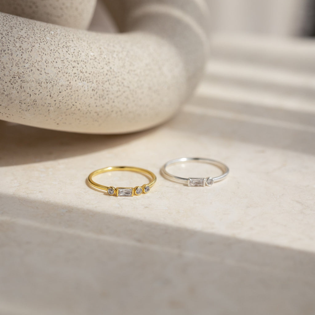 A gold ring and an Initial Morse Code Ring with sparkling stones are displayed on a beige stone surface in natural light, highlighting the elegance of personalized jewelry.