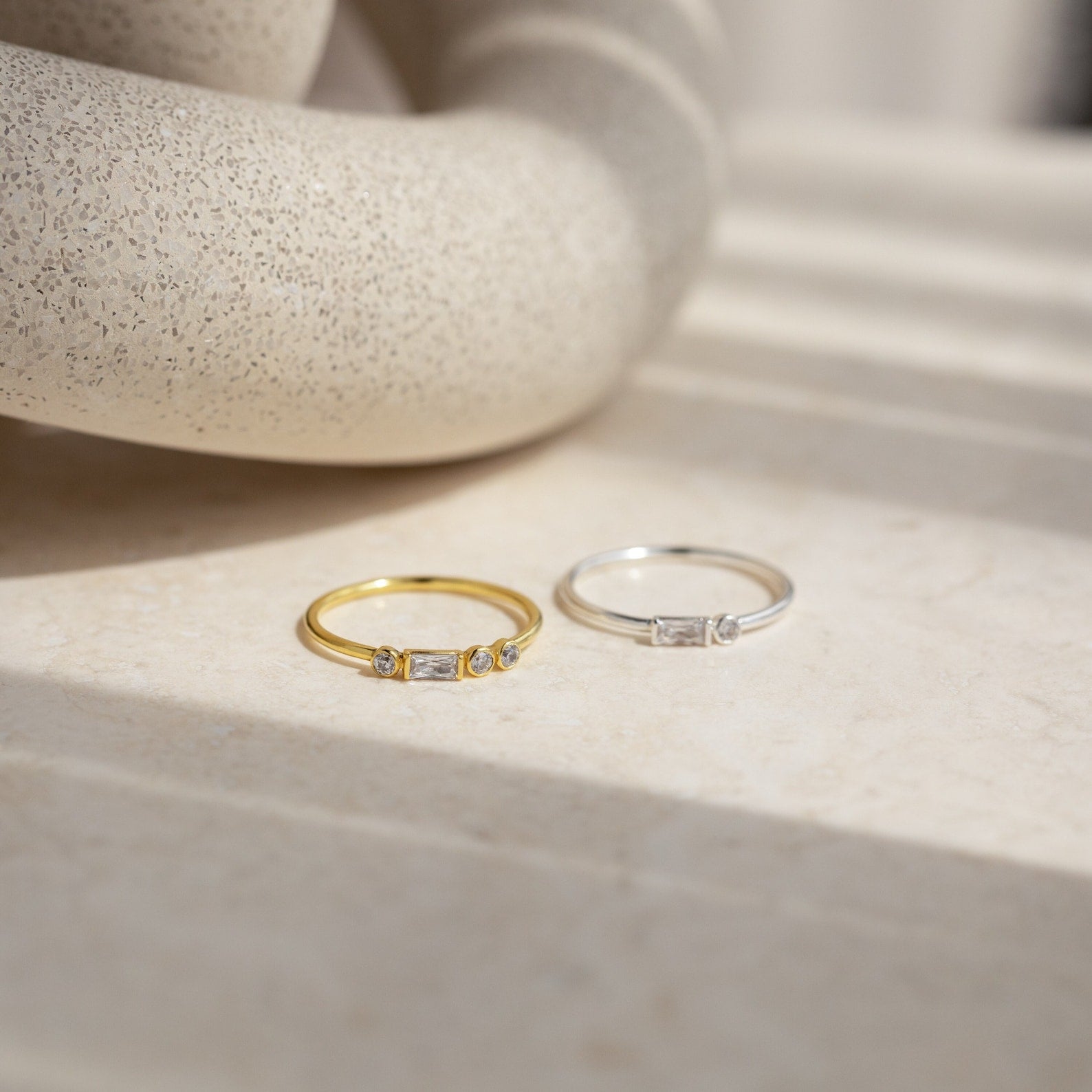 A gold ring and an Initial Morse Code Ring with sparkling stones are displayed on a beige stone surface in natural light, highlighting the elegance of personalized jewelry.