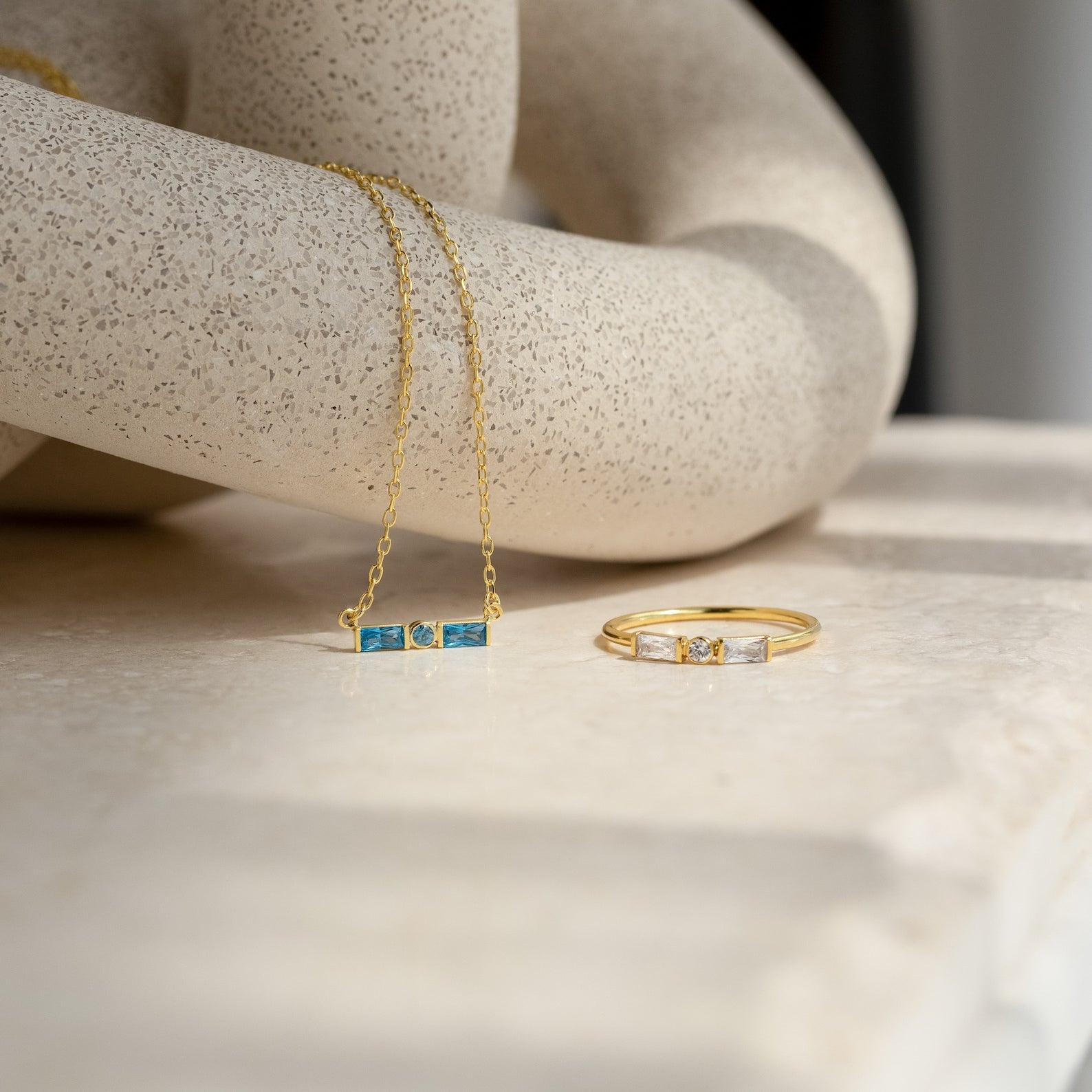 Gold necklace with blue rectangular stones and an Initial Morse Code Ring—personalized gold jewelry with clear stones—displayed on a marble surface.