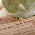 Mushroom Studs—small gold earrings with colorful gemstones shaped like mushrooms—rest on a wooden surface, making them the perfect piece of nature-inspired jewelry.