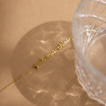 A gold cursive name necklace rests on a beige surface beside a textured glass casting patterns—perfect for minimal jewelry fans or pairing with the Petite Tiny Multiple Name Bracelet for a unique, personalized style.