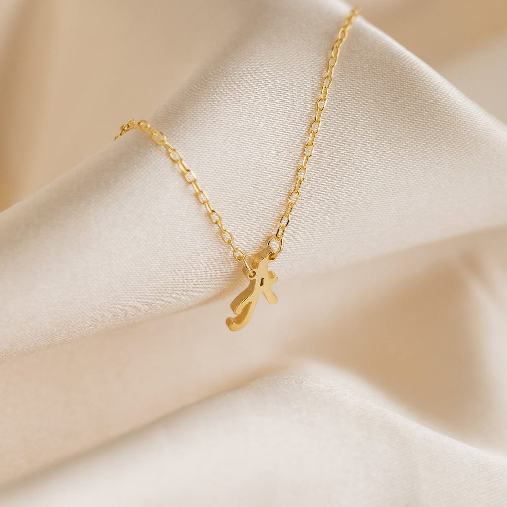 The Stylish Dainty Initial Necklace features a minimalist gold chain with a script letter A pendant, elegantly displayed on cream fabric—an ideal personalized accessory for everyday style.