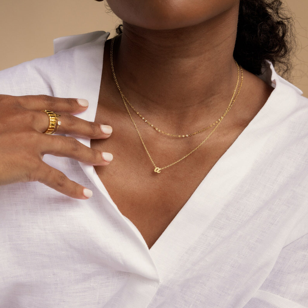 A woman in a white blouse wears layered gold necklaces, including a dainty pendant and the Mellow Dainty Initial Necklace, along with a gold ring on her finger.