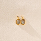 Moonstone Drop Earrings feature gold-framed oval moonstones with small round accents, displayed on a textured beige surface—a perfect minimalist accessory and thoughtful graduation gift.
