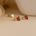 Garnet Heart Studs with heart-shaped garnet stones and clear gems rest elegantly on a beige surface—perfect earrings for any occasion.