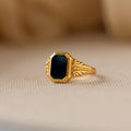 The Vintage Black Signet Ring features ornate gold detailing and a bold, black octagonal gemstone, evoking classic vintage style and displayed on a beige surface.