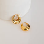 The Katrina Ribbed Hoops, featuring a chunky ribbed texture and layered crescent design, rest on a smooth white surface, highlighting their modern style.