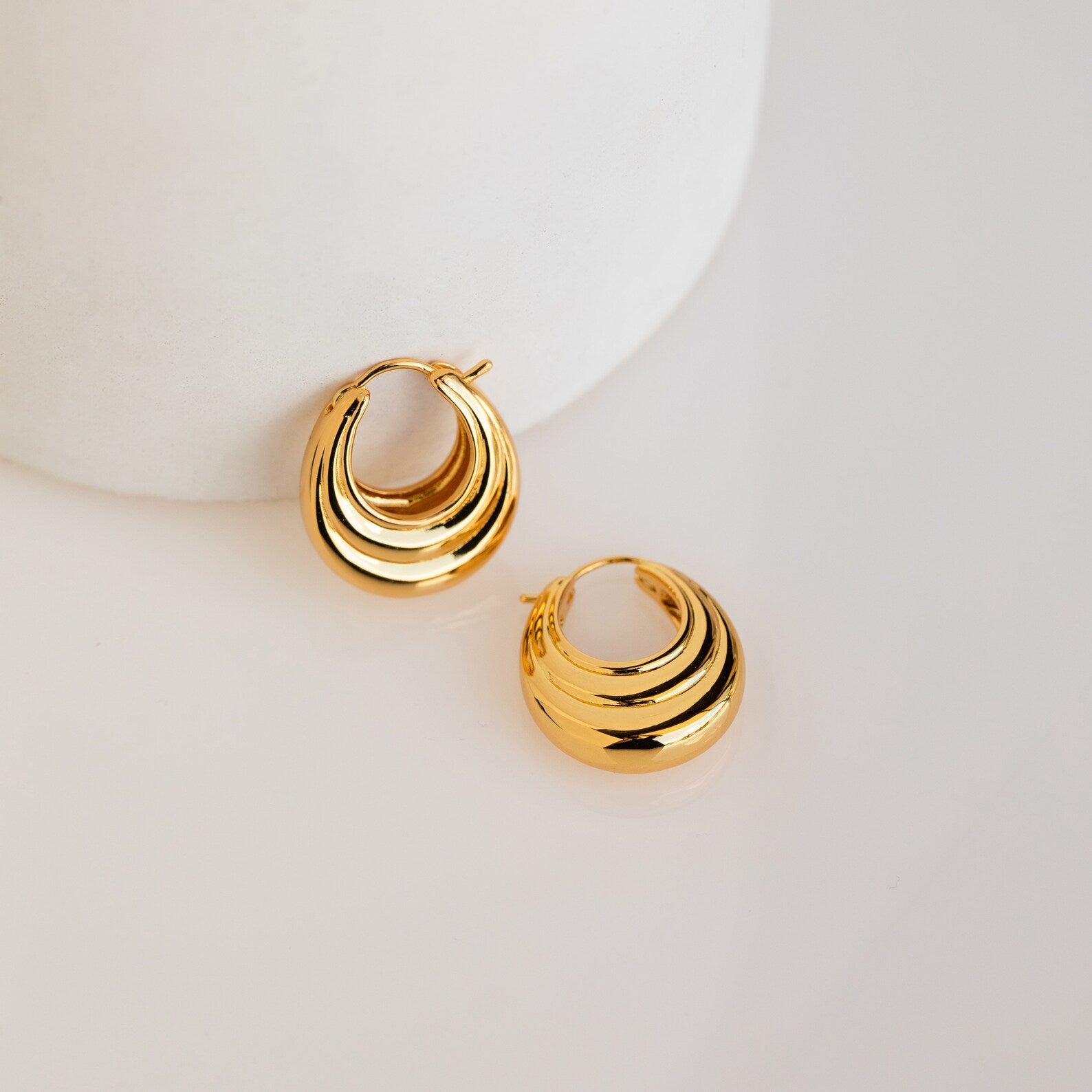 The Katrina Ribbed Hoops, featuring a chunky ribbed texture and layered crescent design, rest on a smooth white surface, highlighting their modern style.