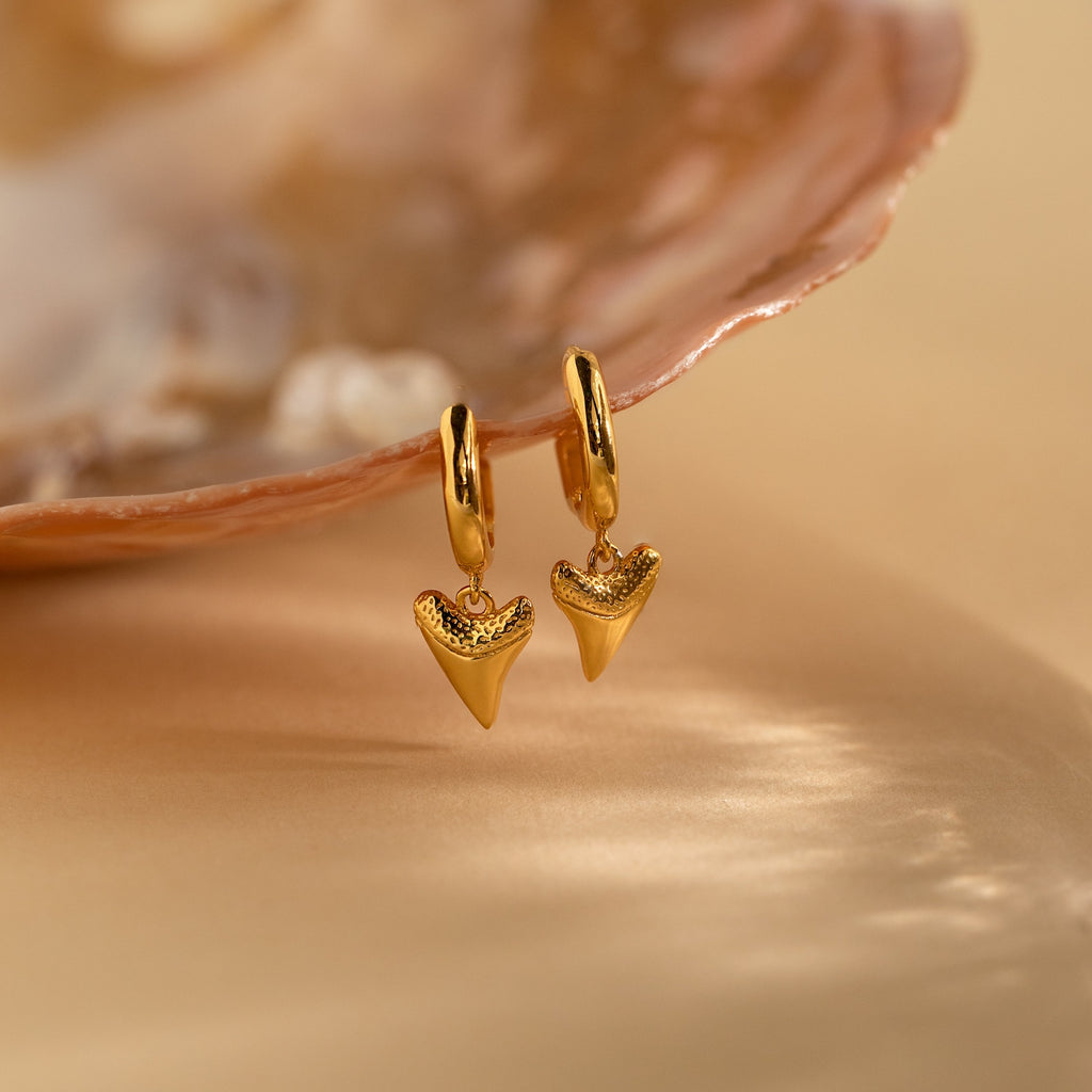 Shark Tooth Huggies: Gold huggie hoop earrings featuring small shark tooth pendants, shown next to an open seashell—an eye-catching piece of beach-inspired jewelry.