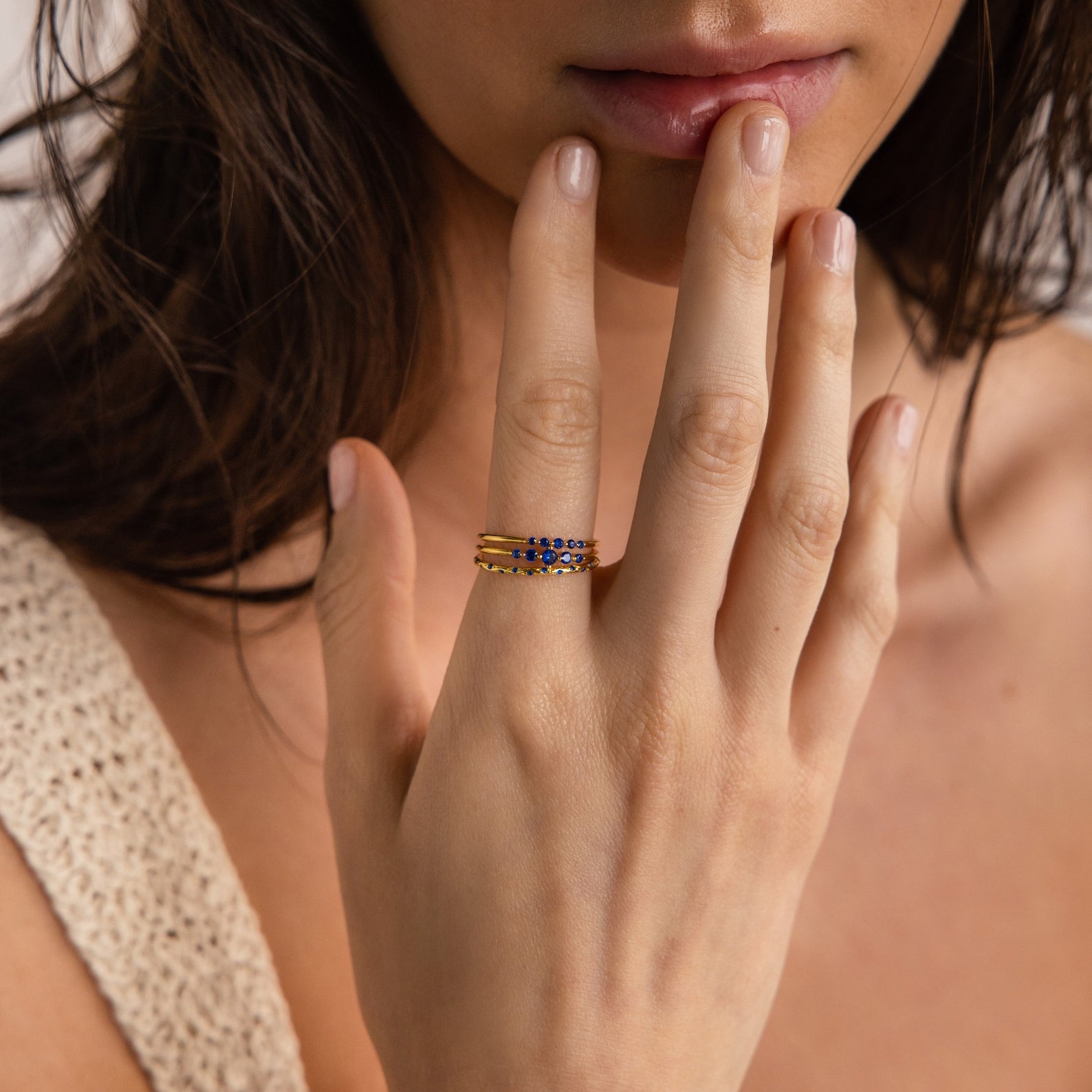 A woman wears the Sapphire Stacking Ring Set—gold rings with blue sapphires—as she touches her lips against a neutral background.