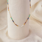 The Oasis Rainbow Beaded Necklace, featuring small pearls and colorful beads, drapes elegantly over a textured white vase—making it the perfect boho jewelry or summer accessory.