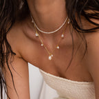 A woman wears the Baroque Pearl Station Necklace layered with minimalist necklaces and a cream strapless textured top, highlighting her shoulders and collarbone.