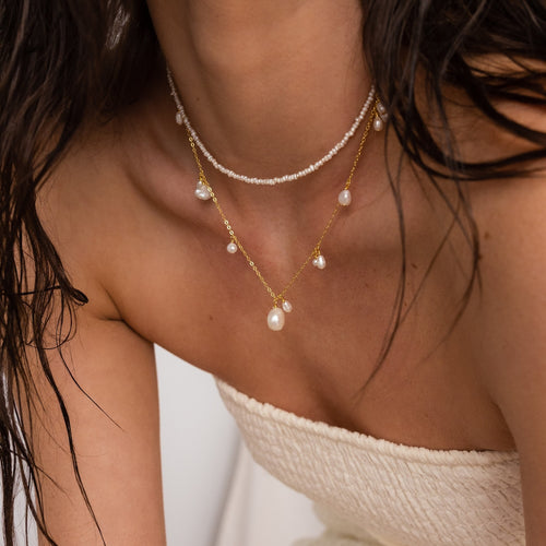 A woman wears the Baroque Pearl Station Necklace layered with minimalist necklaces and a cream strapless textured top, highlighting her shoulders and collarbone.