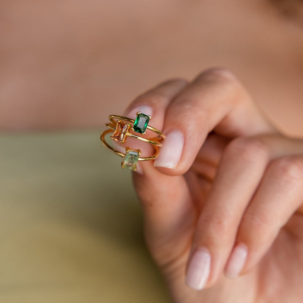 A hand holds three Emerald Cut Solitaire Rings, each set with a rectangular green, yellow, or peach gemstone—ideal as elegant birthstone rings or striking statement pieces.