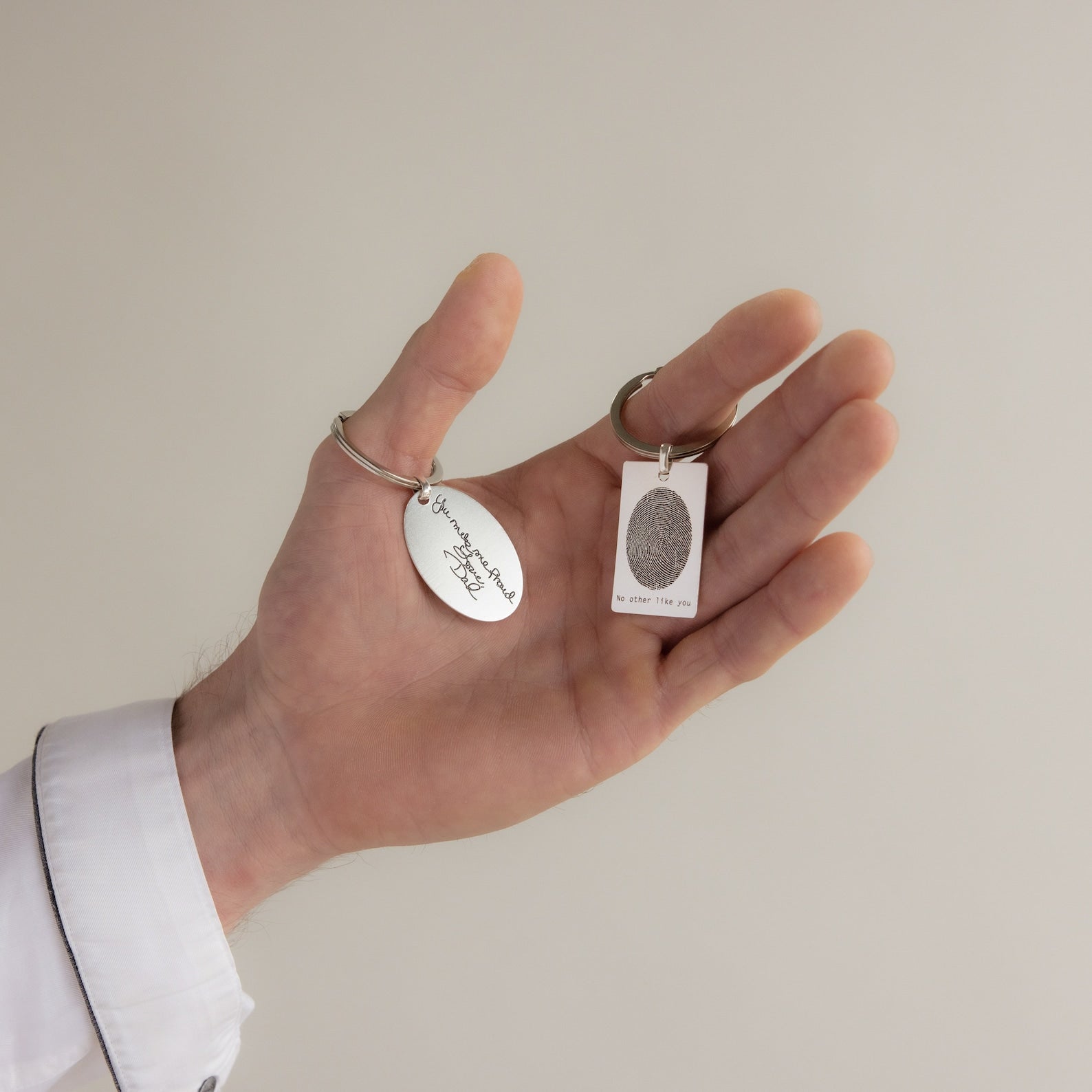 A hand holding two silver keychains, including the Fingerprint Rectangle Keychain with custom handwriting and fingerprint— a unique personalized keepsake for someone special.