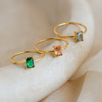 Three gold rings with emerald-cut green, peach, and blue stones are displayed on a light textured surface; one is the Citrine Emerald Ring, perfect for those with a November birthstone.