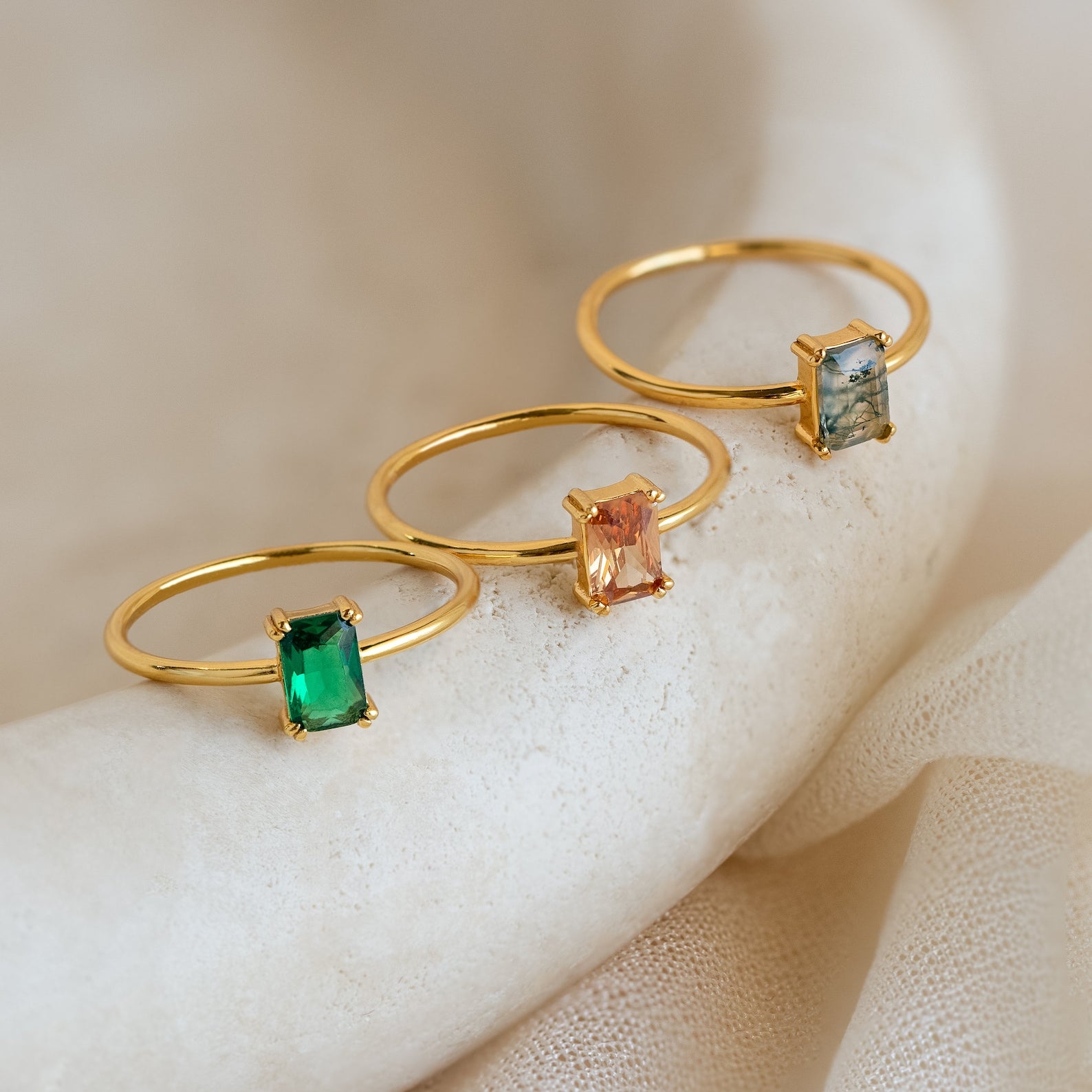 Three Emerald Cut Solitaire Rings with green, peach, and clear stones are displayed on a textured surface. Perfect for stacking or wearing alone, these rings make a stunning statement.