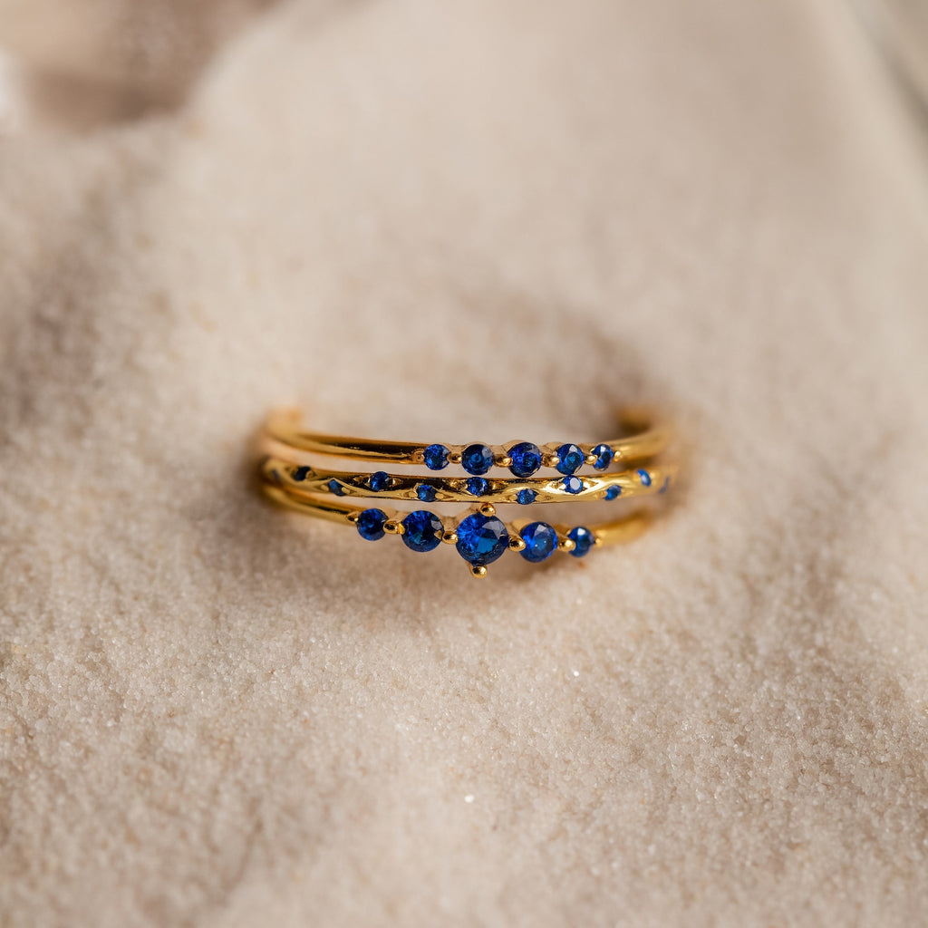 The Sapphire Stacking Ring Set, featuring blue sapphires set in gold, rests elegantly on a white surface.