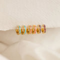 Six Pastel Opal Inlay Huggies—small gold hoops accented with colorful enamel—are displayed on a textured cream fabric background.