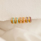 Six Pastel Opal Inlay Huggies—small gold hoops accented with colorful enamel—are displayed on a textured cream fabric background.
