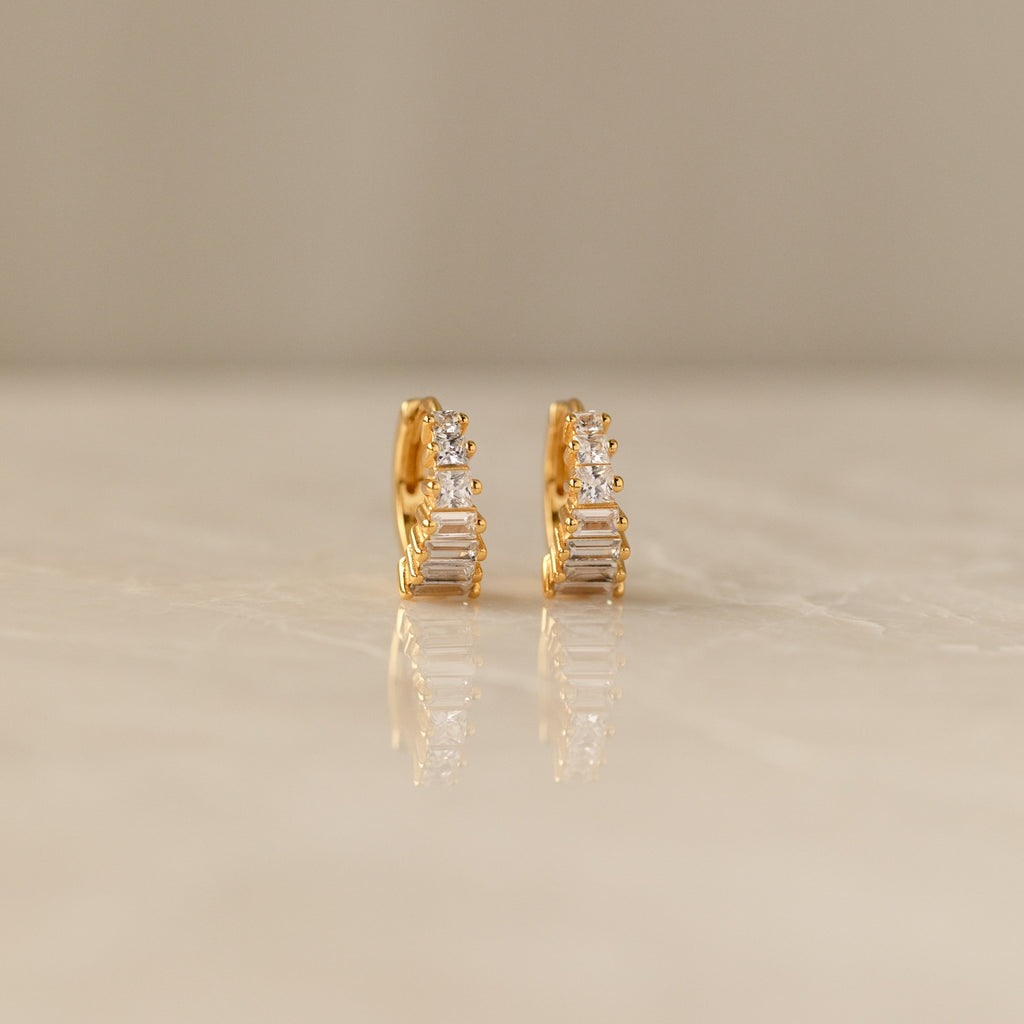 Close-up of a pair of gold Art Deco Huggie earrings on a marble surface, featuring a symmetrical arrangement of baguette and round-cut diamonds for a tiered, structured look.