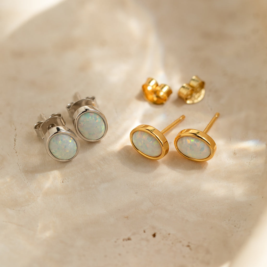 Two pairs of Oval Opal Studs, in silver and gold, are displayed on a light marble surface, each showcasing a luminous white opal stone.