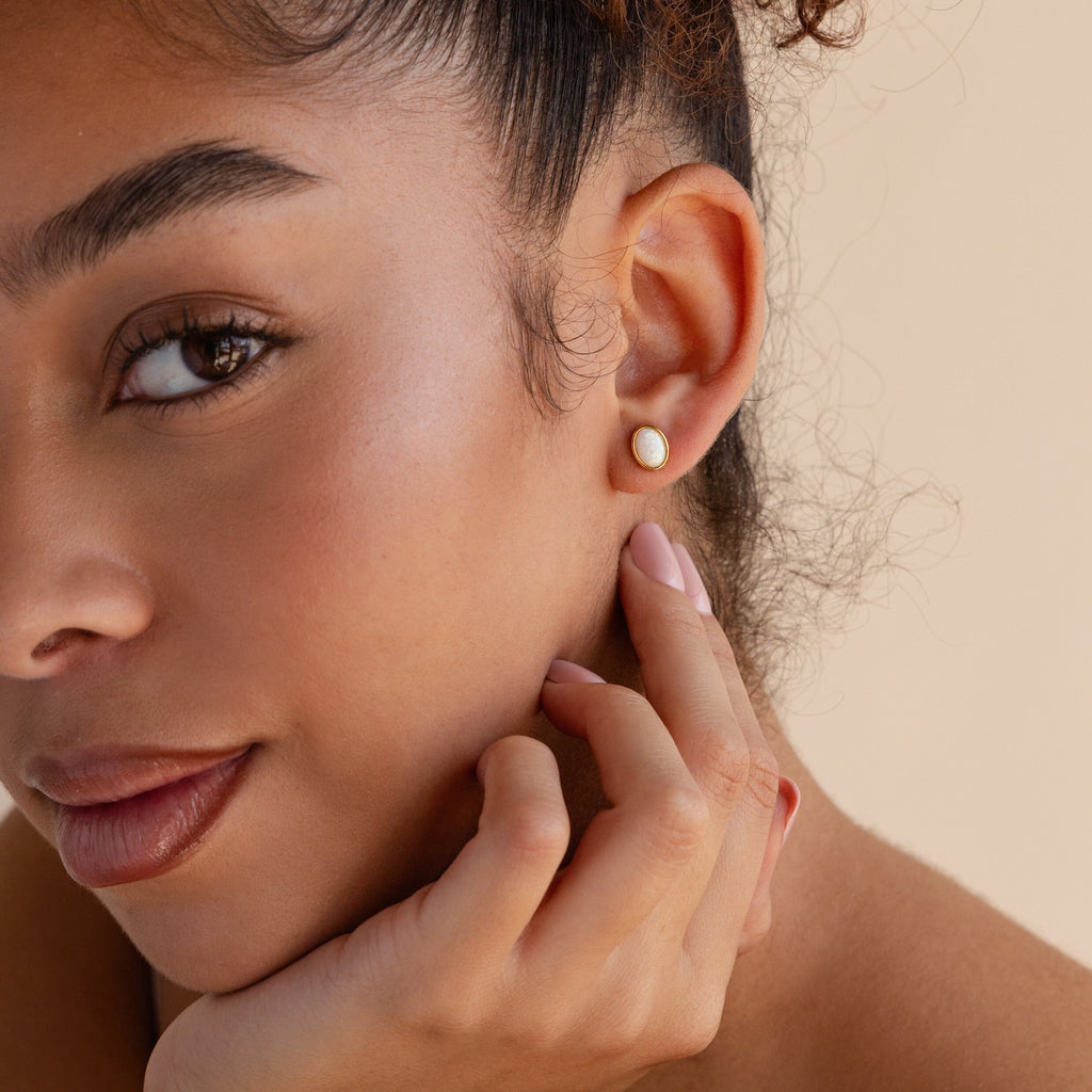 A woman with natural makeup touches her face, showcasing the elegant Oval Opal Studs—refined stud earrings featuring a subtle white opal stone—alongside her manicured nails.