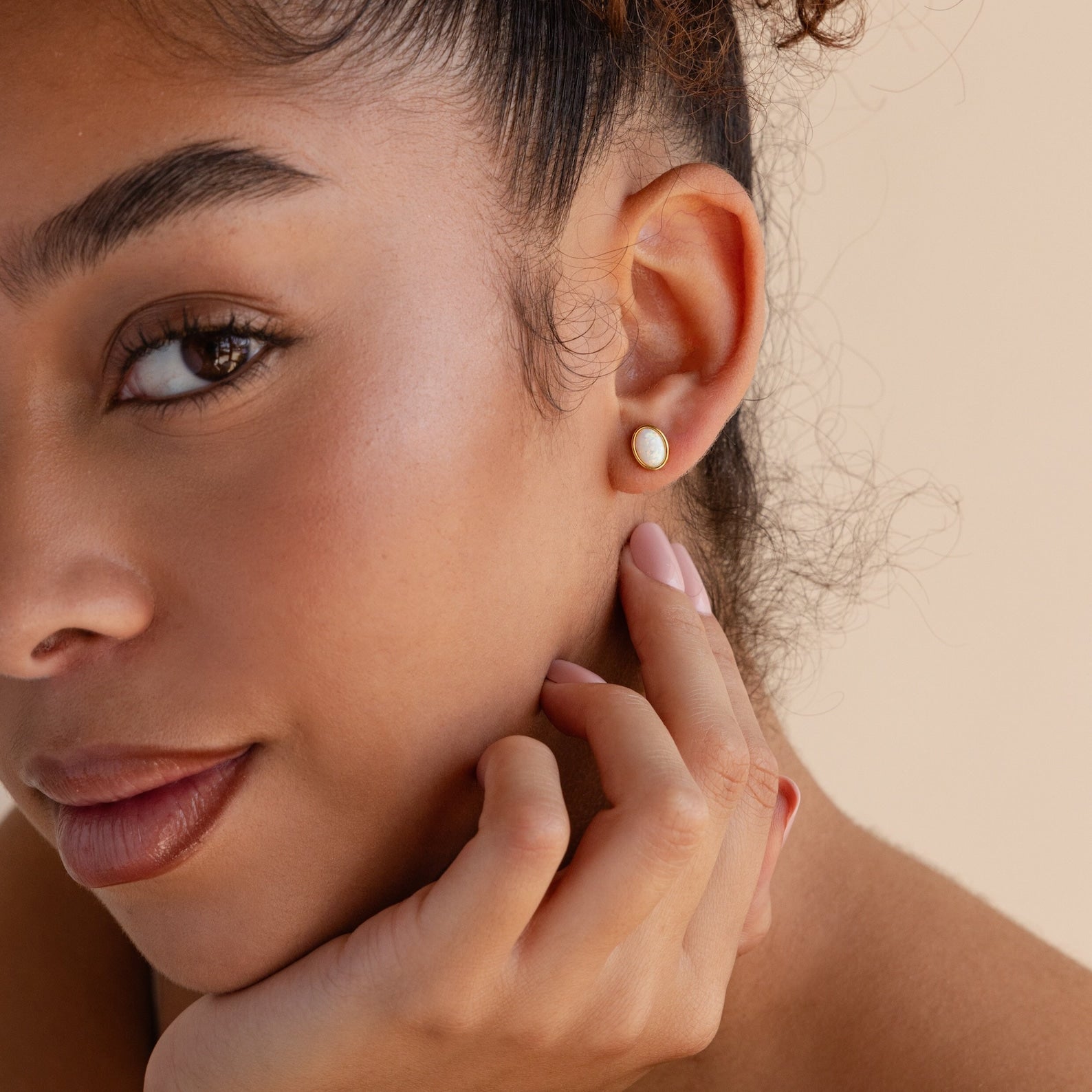 A woman with natural makeup touches her face, showcasing the elegant Oval Opal Studs—refined stud earrings featuring a subtle white opal stone—alongside her manicured nails.
