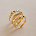 Three Thin Name Signet Rings, each gold, engraved with a name and fingerprint, set with a small gemstone, displayed on a light background.