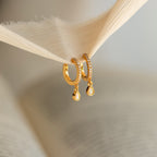 Opal Drop Huggies—gold huggie hoops with delicate opal drops—are softly lit as they hang from a piece of beige fabric.