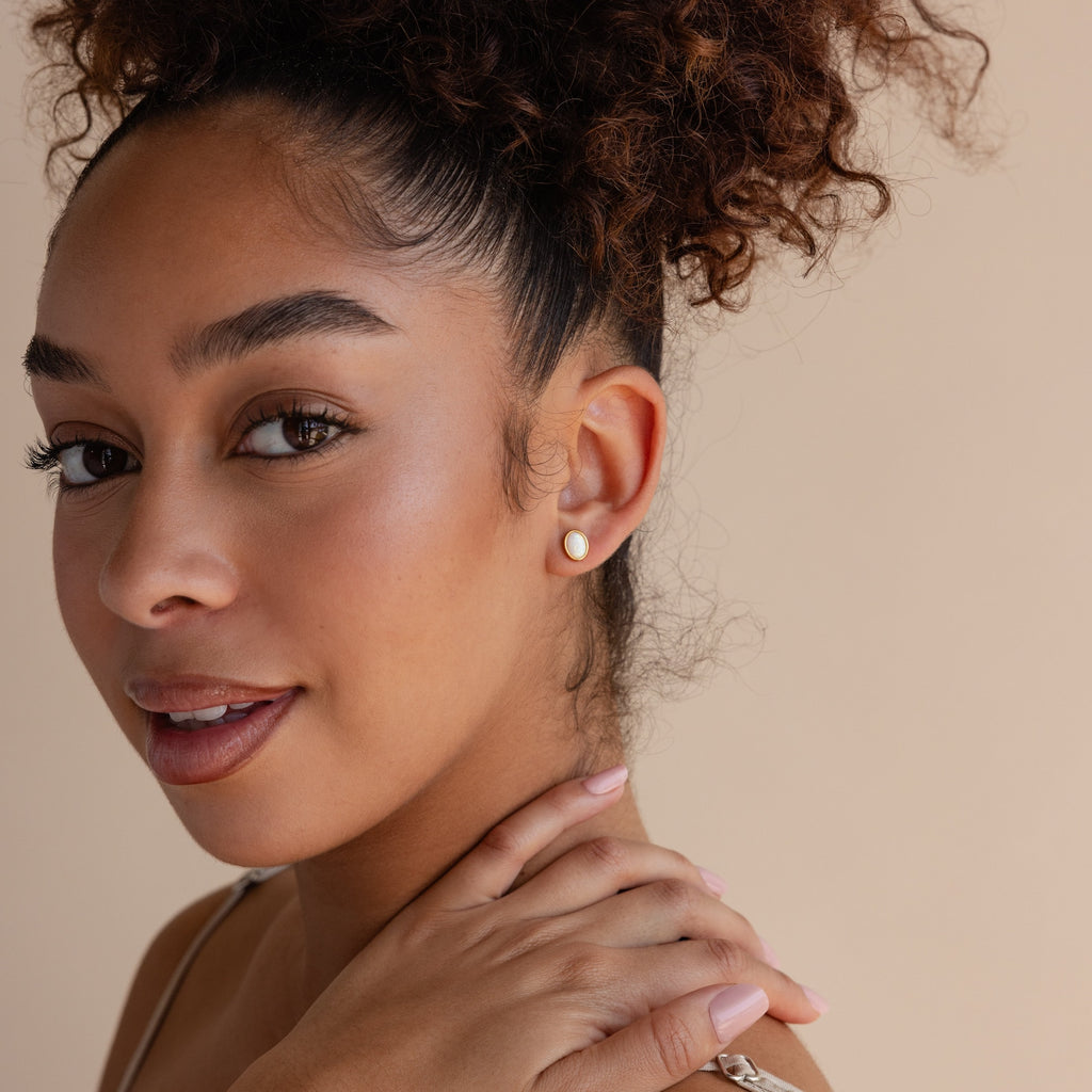 A woman with curly hair smiles warmly, her hand on her shoulder, showcasing elegant Oval Opal Studs against a neutral background.