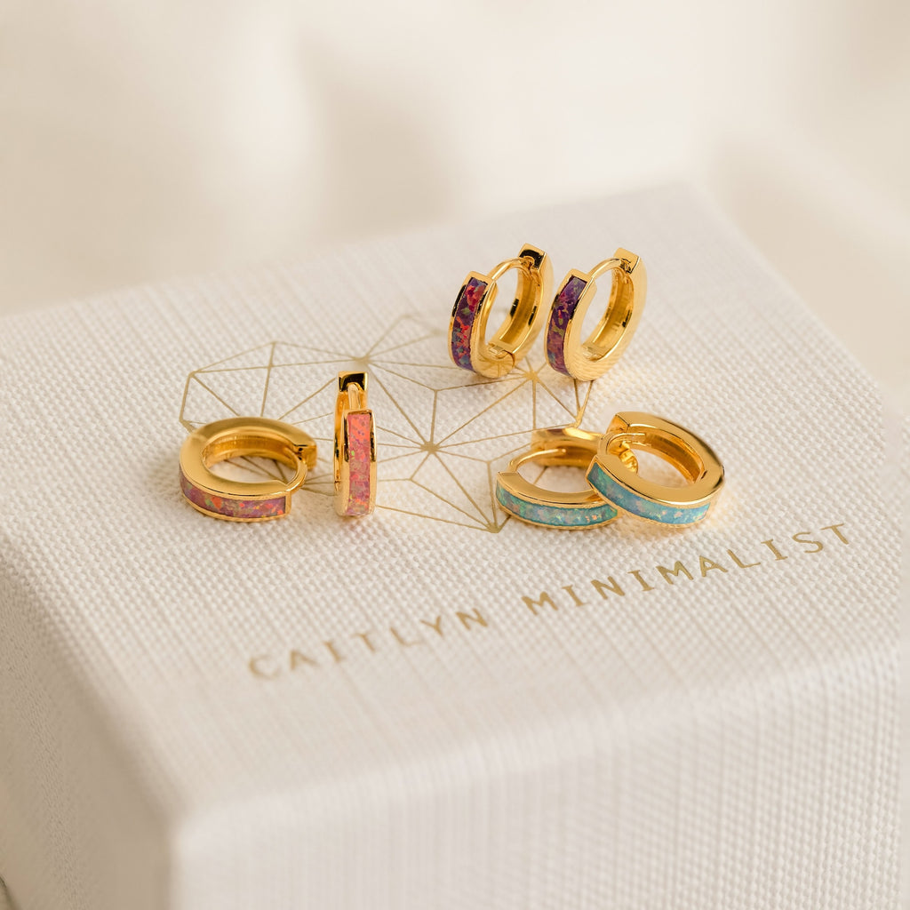 Five pairs of small gold hoop earrings, including Ocean Opal Inlay Huggies with colorful inlays, displayed on a textured box labeled Caitlyn Minimalist—perfect as chic wedding jewelry or everyday accessories.