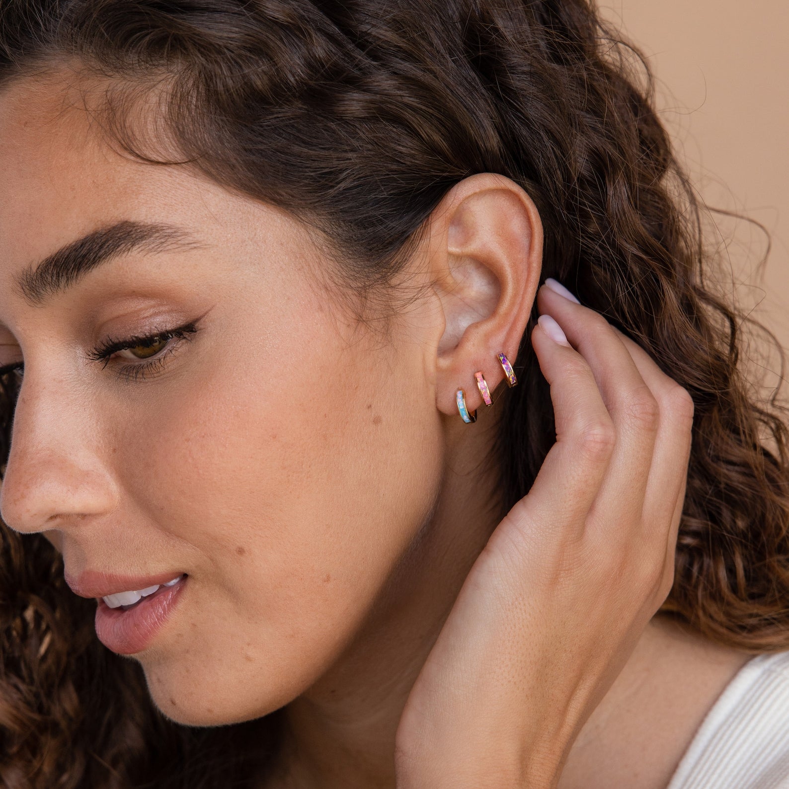 A woman with curly hair wears colorful small hoop earrings, including Ocean Opal Inlay Huggies, as she touches her ear and smiles softly.