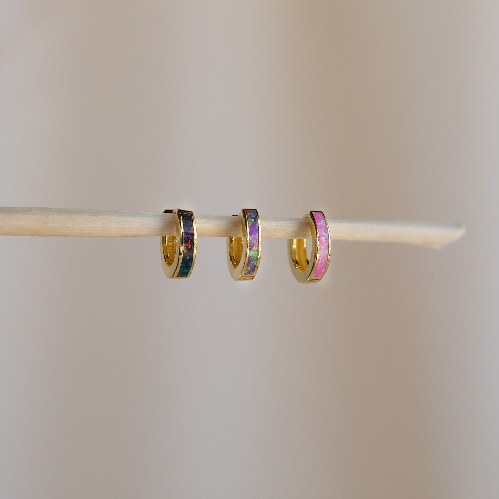 Three small minimalist hoop earrings hang on a thin wooden stick, including the Lavender Opal Inlay Huggies, which add a subtle purple opal shimmer against a plain background.