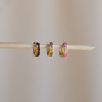 Three small, colorful huggie hoop earrings hang on a thin stick against a neutral background, evoking the vibrant style of Black Opal Inlay Huggies.