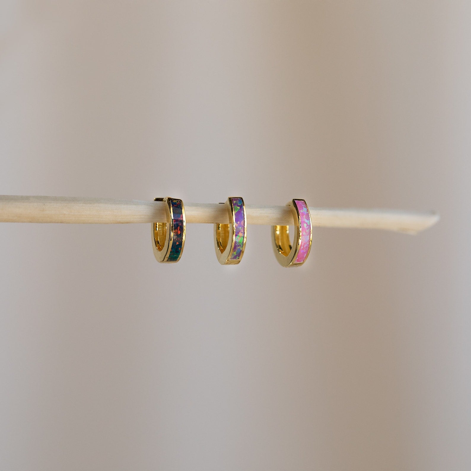 Three small, colorful huggie hoop earrings hang on a thin stick against a neutral background, evoking the vibrant style of Black Opal Inlay Huggies.