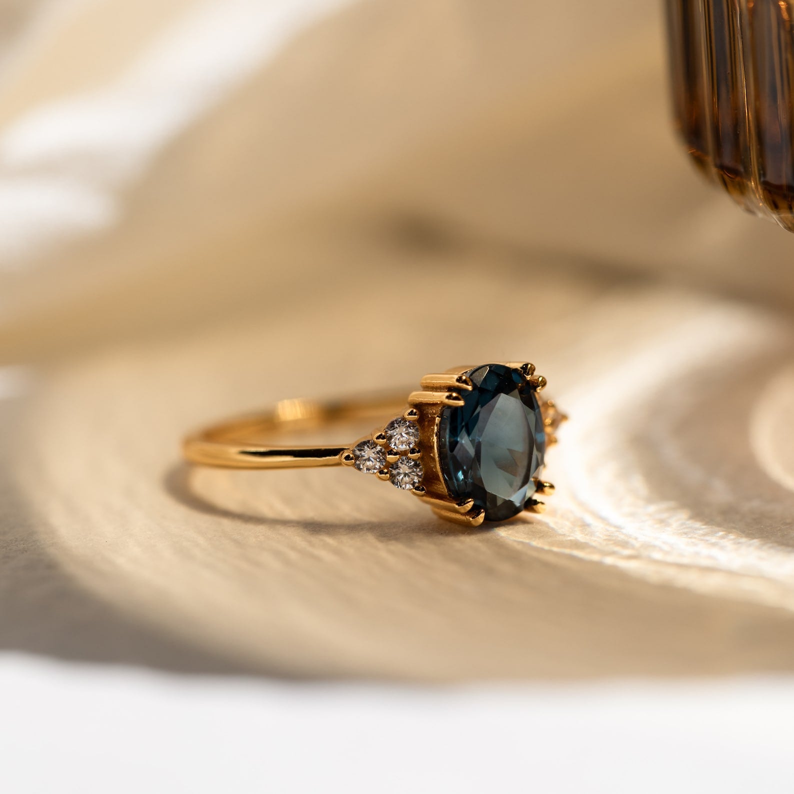 Close-up of the Elizabeth ring on a soft fabric surface, showcasing the oval-cut blue topaz and side stones.