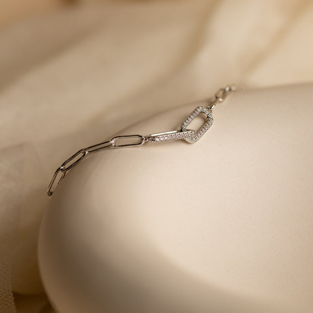 The Pave Link Chain Bracelet features delicate geometric links in silver, resting on a cream-colored surface—a minimalist and understated statement piece.